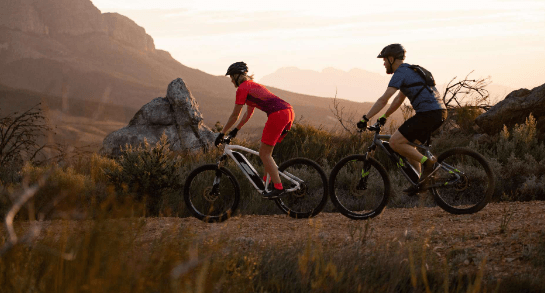 Two riders on electric bikes mountain biking on a dirt trail at sunset, side view