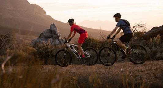 Two riders on electric bikes mountain biking on a dirt trail at sunset, side view
